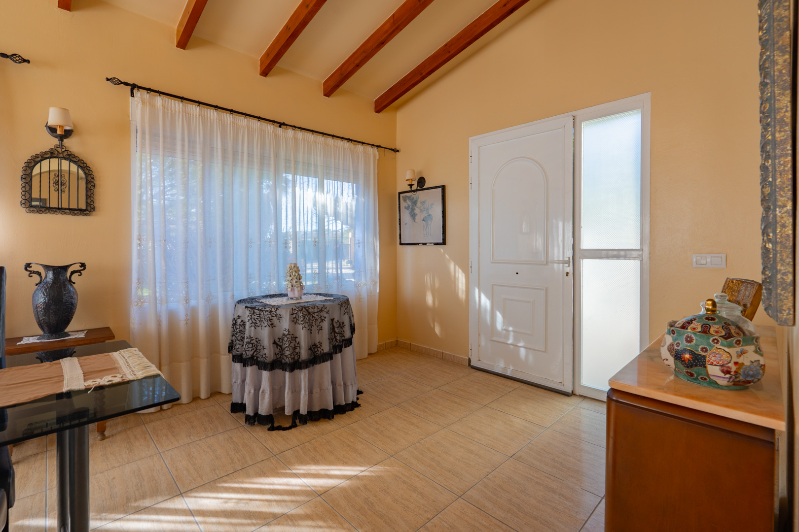 Vestíbulo de entrada con techos altos de vigas de madera, suelos cerámicos y una puerta principal blanca con panel lateral de cristal translúcido.