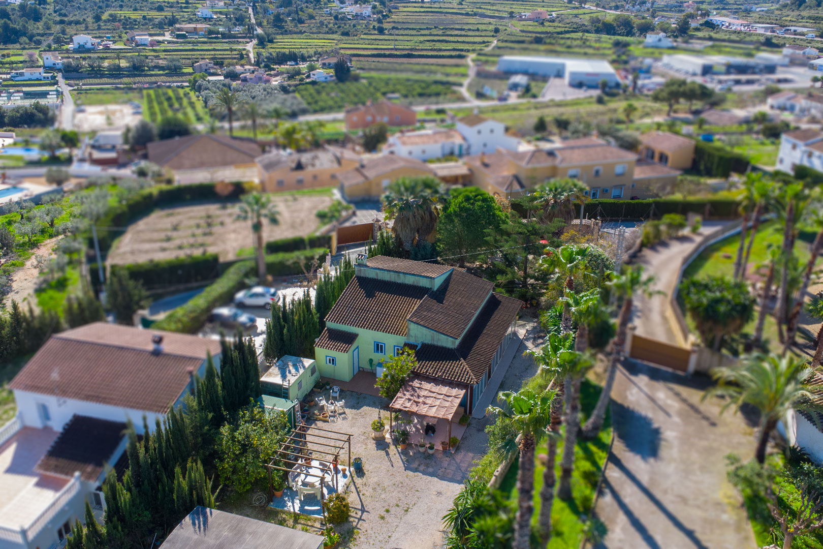 Vista aérea de la finca tradicional con fachada verde, destacando su amplia parcela de grava, pérgolas de madera y palmeras que bordean el acceso principal.