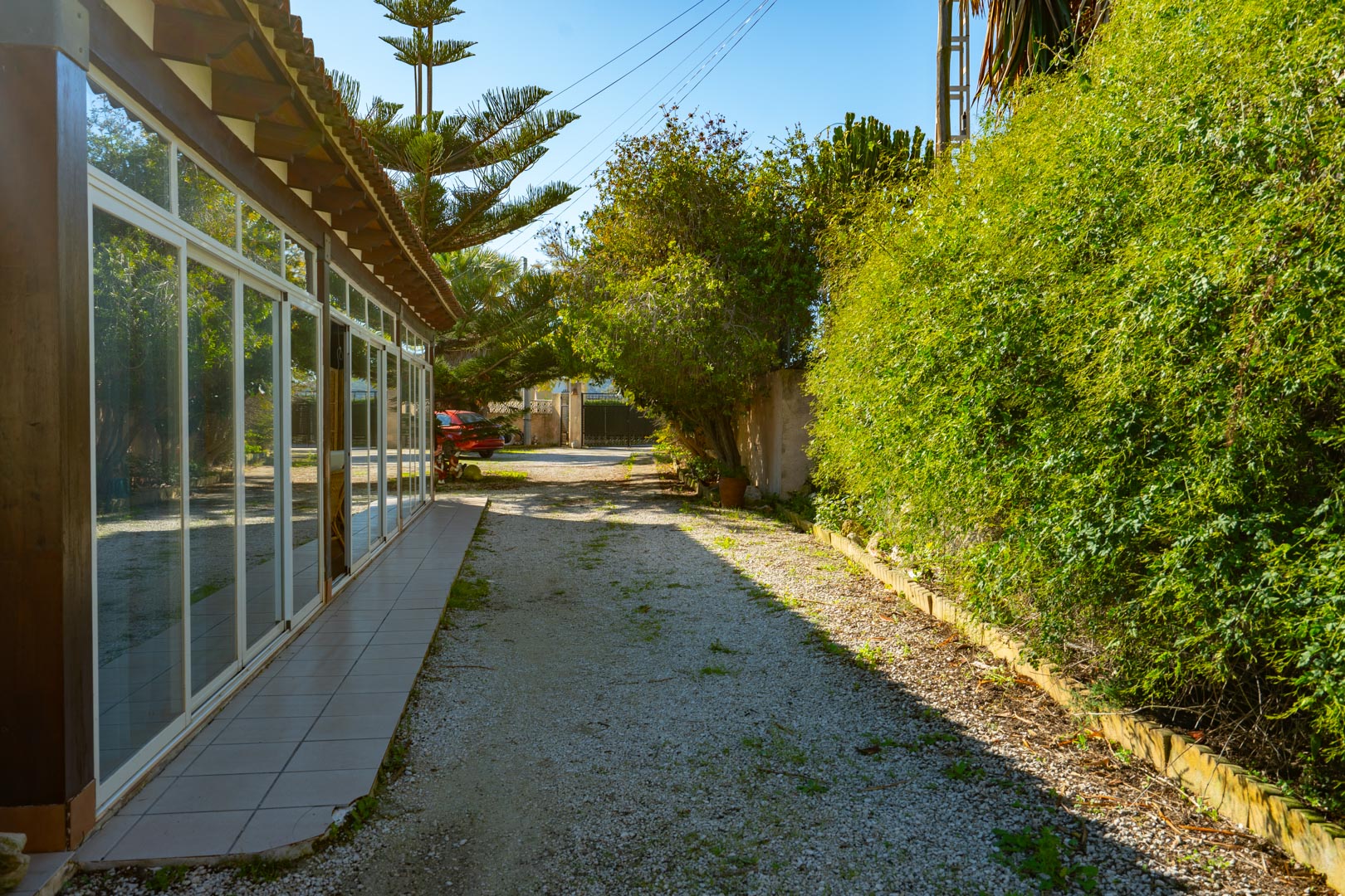 Vista exterior de la naya acristalada con vigas de madera, situada junto a un camino de grava rodeado de vegetación mediterránea madura.
