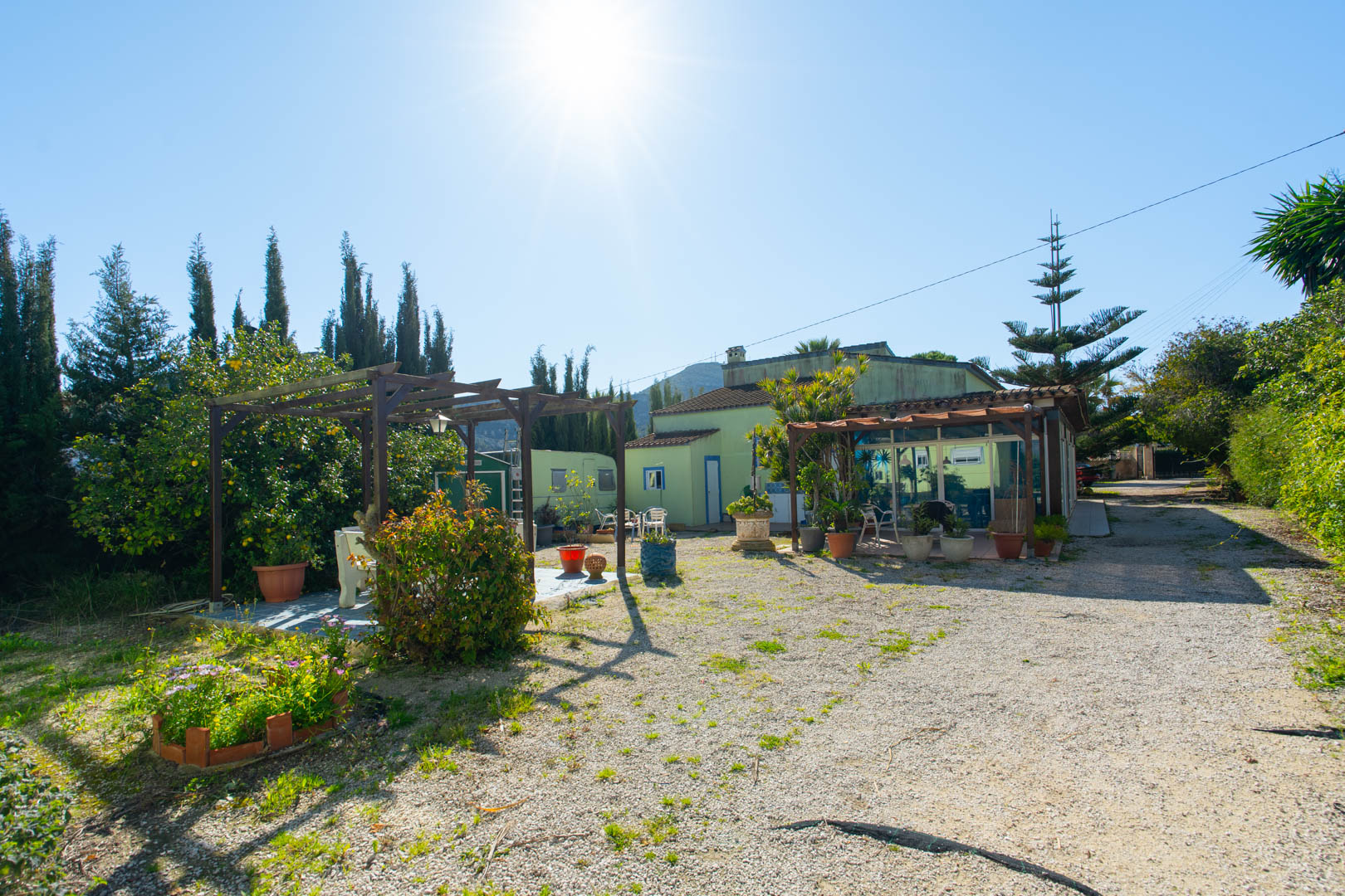 Vista exterior del jardín rústico con pérgola de madera, zona de estar y la fachada de la vivienda con naya acristalada y camino de grava.