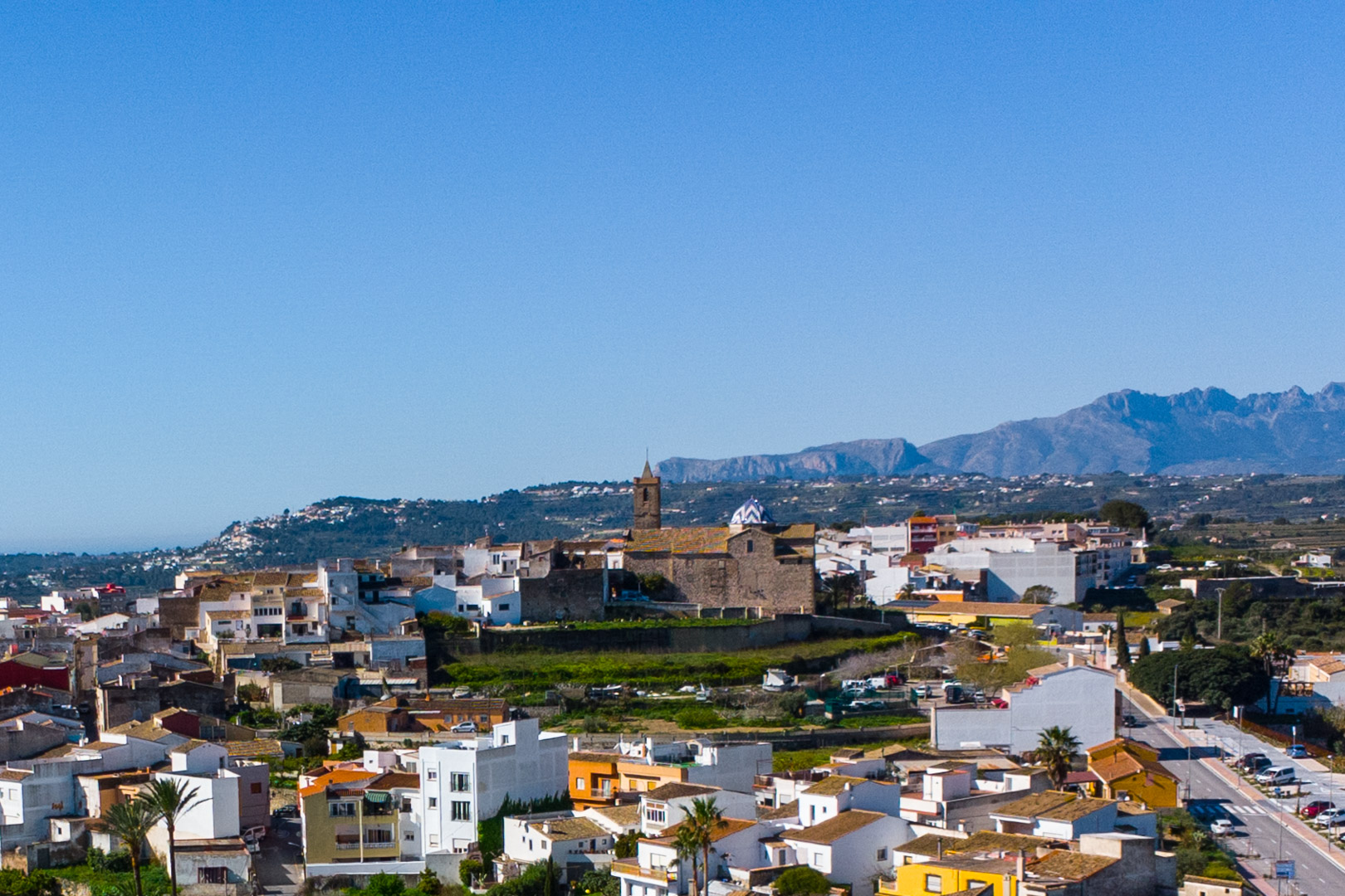Vista panorámica del casco urbano de Benitachell con la iglesia de cúpula azul y el macizo del Montgó al fondo bajo un cielo despejado.
