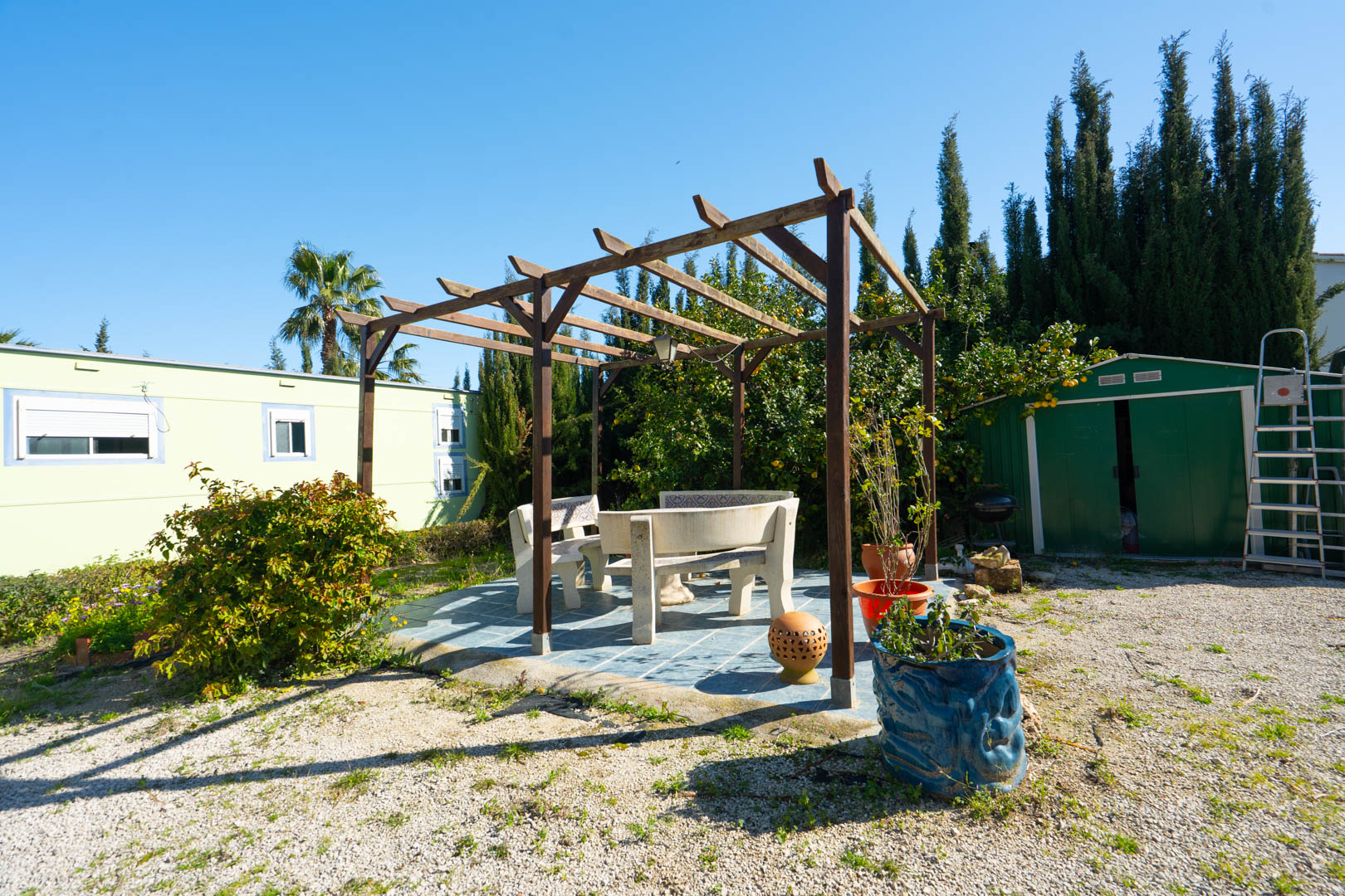 Zona de estar exterior con pérgola de madera y bancos de piedra sobre suelo cerámico, rodeada de vegetación mediterránea y dependencias auxiliares.