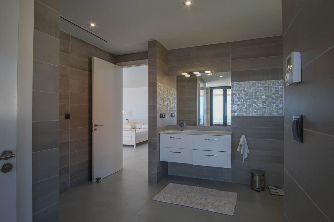 Baño moderno con azulejos grises de gran formato, lavabo suspendido blanco y detalle de mosaico. La puerta abierta conecta con el dormitorio y ofrece vistas al mar.