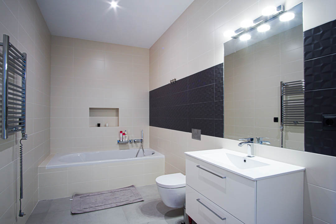 Baño moderno con bañera encastrada, sanitario suspendido y mueble de lavabo blanco. Paredes alicatadas en tonos crema con una franja decorativa texturizada en gris oscuro.
