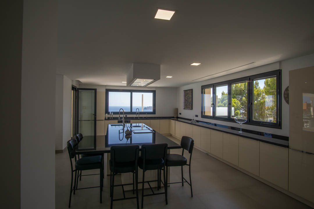 Cocina moderna con isla central de granito negro, armarios minimalistas en tono crema y amplios ventanales con vistas directas al mar Mediterráneo y pinos.