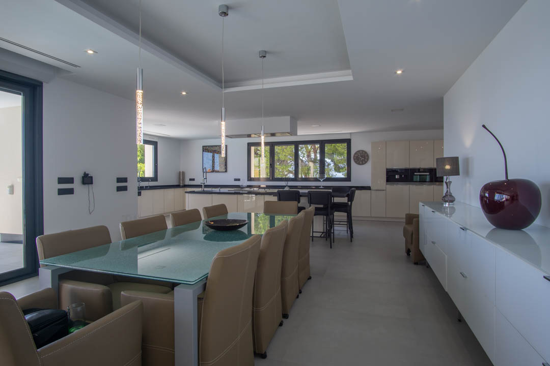 Comedor de concepto abierto con mesa de cristal y sillas de cuero, integrado con una cocina moderna de encimeras oscuras e iluminación empotrada.