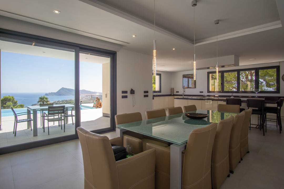 Comedor diáfano con mesa de cristal y sillas de cuero, integrado con cocina moderna y acceso a terraza con vistas a la bahía de Altea y piscina.