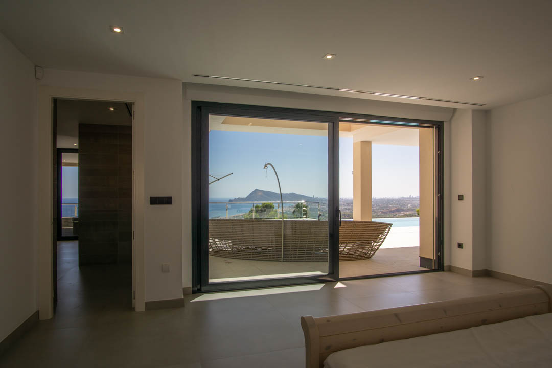 Dormitorio con acceso directo a la terraza mediante ventanales de suelo a techo, ofreciendo vistas despejadas a la bahía de Altea y la montaña.