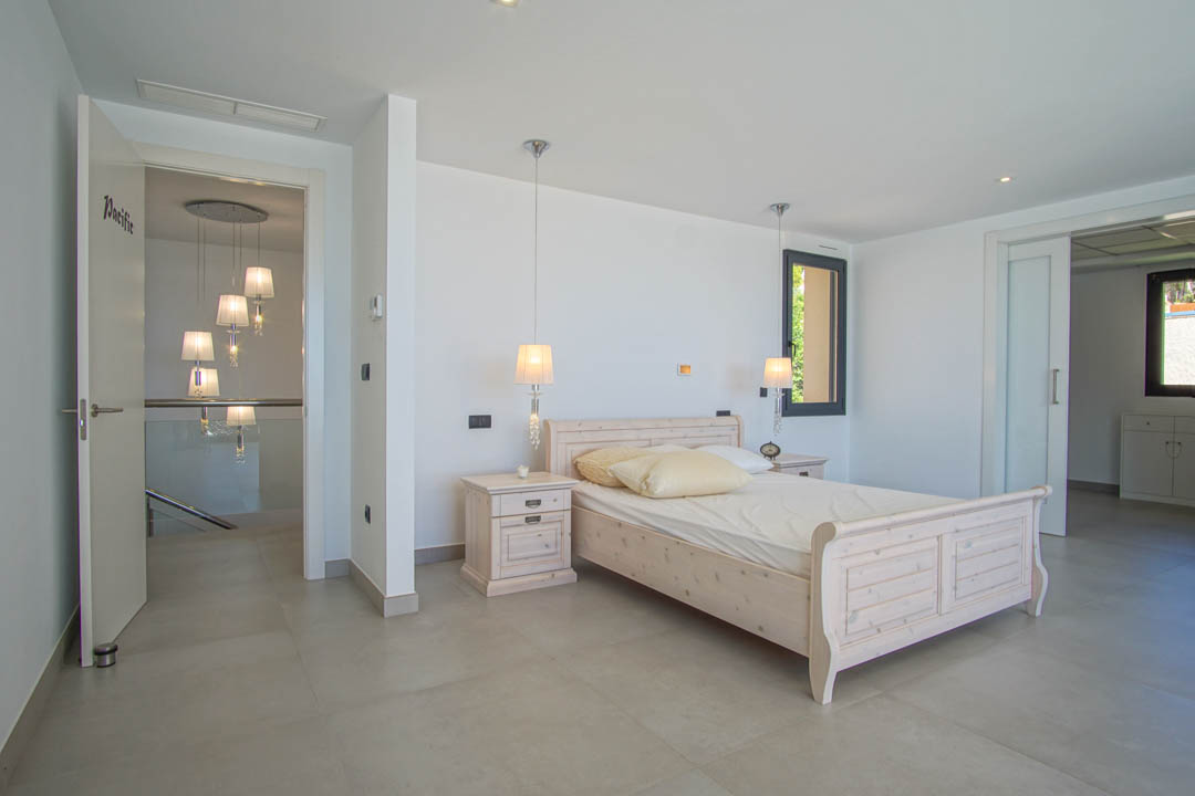 Dormitorio luminoso con cama de madera clara, mesitas a juego y lámparas colgantes. Incluye suelos cerámicos grises y acceso a una zona de vestidor mediante puerta corredera.