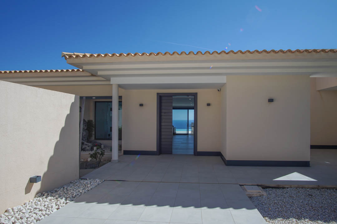 Entrada principal de diseño moderno con puerta gris, suelos de gres porcelánico y vistas directas al mar a través del vestíbulo bajo un cielo despejado.