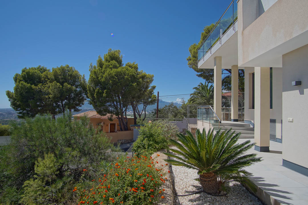 Exterior de la villa con fachada blanca, columnas estructurales y jardines mediterráneos con palmeras y flores. La terraza superior cuenta con barandillas de cristal.