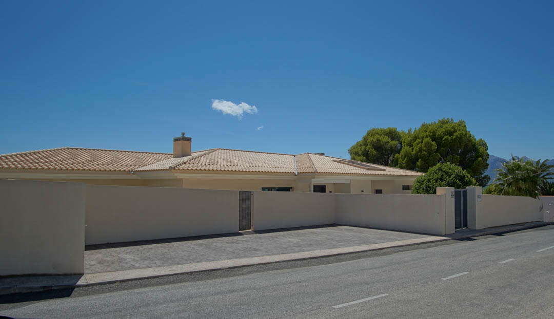 Fachada exterior con muro perimetral, zona de aparcamiento pavimentada para cuatro vehículos y tejado de teja clara bajo un cielo despejado.