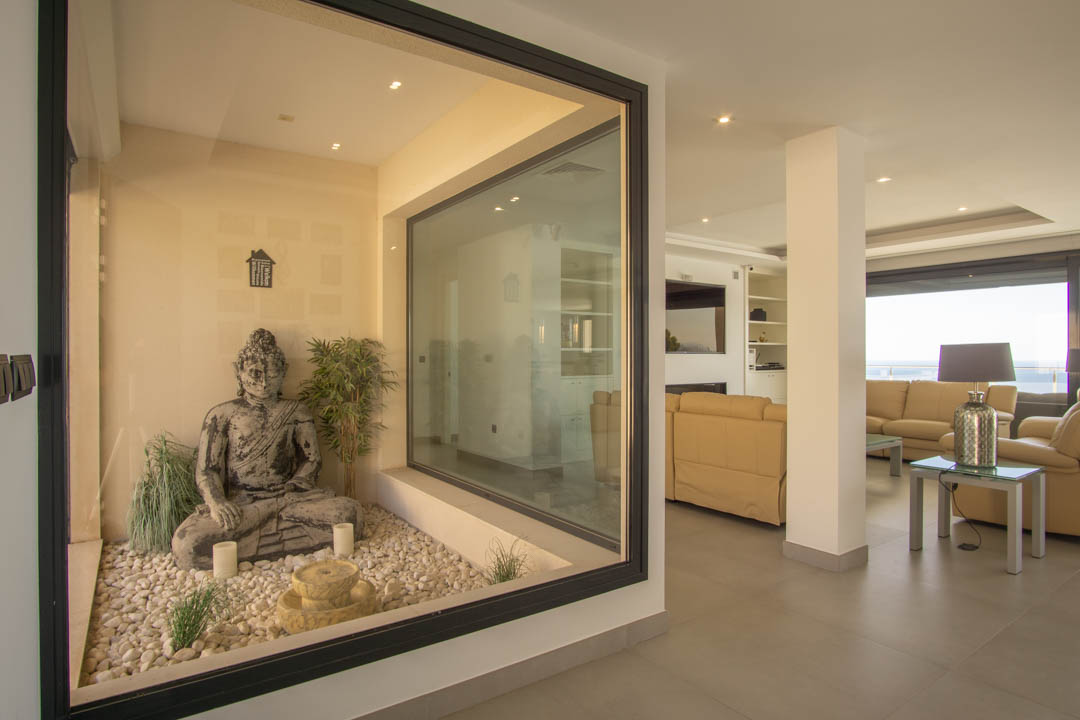 Jardín interior zen acristalado con estatua de Buda y piedras blancas, conectado visualmente con el amplio salón de tonos neutros e iluminación empotrada.