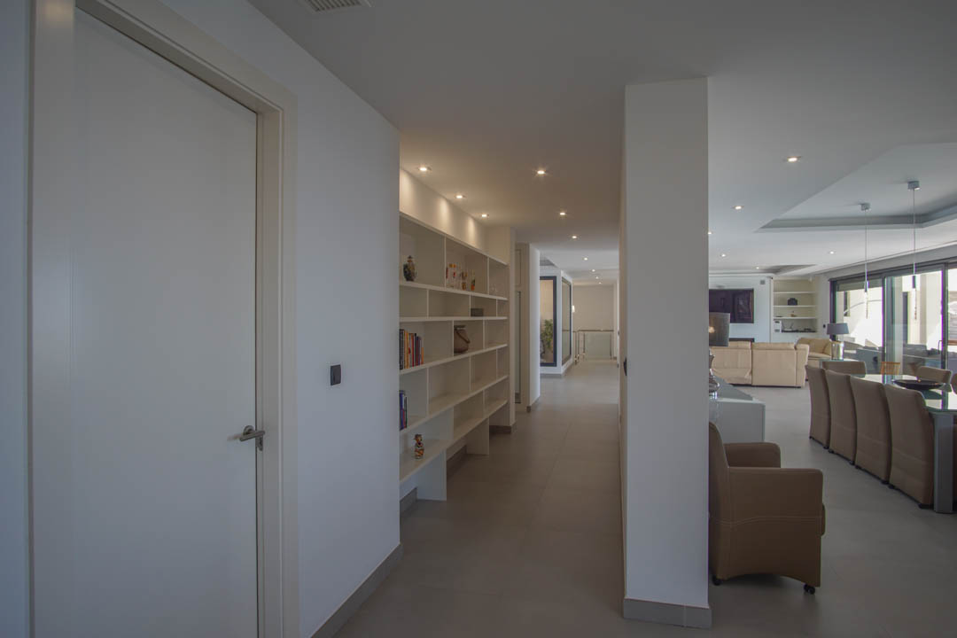 Pasillo de distribución con estantería blanca integrada y focos empotrados que conecta con el amplio salón-comedor de planta abierta y grandes ventanales.