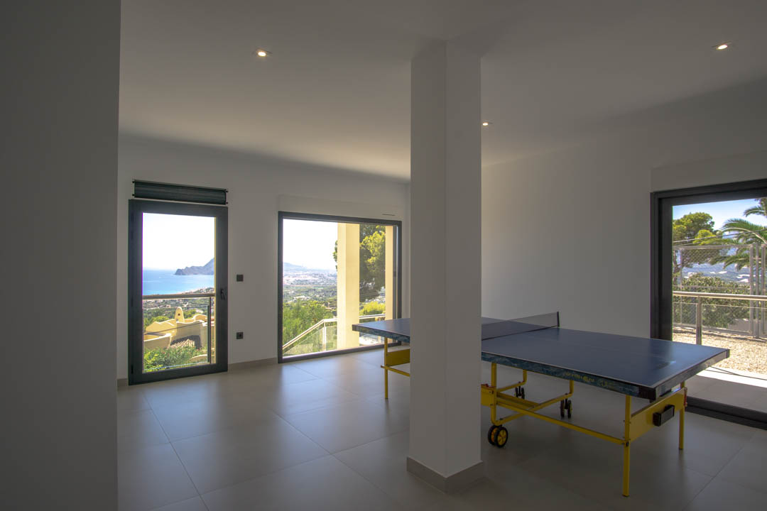 Sala de juegos con mesa de ping-pong, suelos cerámicos grises y amplios ventanales con vistas panorámicas a la costa de Altea y el mar Mediterráneo.