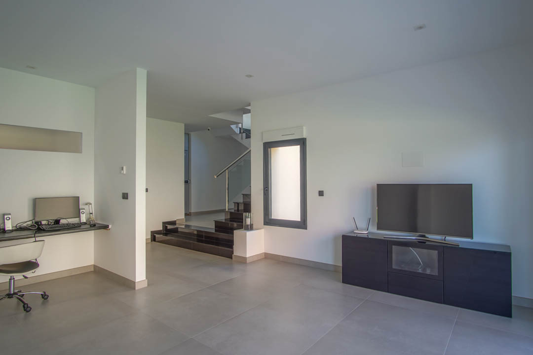 Salón de planta baja con zona de despacho integrada, mueble de televisión minimalista y acceso a la escalera de peldaños oscuros con barandilla de cristal.