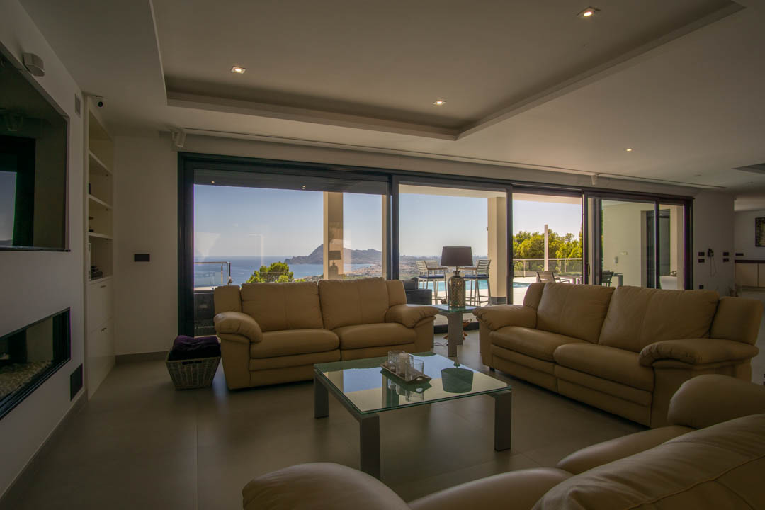 Salón espacioso con sofás de cuero beige y grandes ventanales correderos que ofrecen vistas panorámicas a la bahía de Altea. Incluye chimenea integrada e iluminación empotrada en el techo.