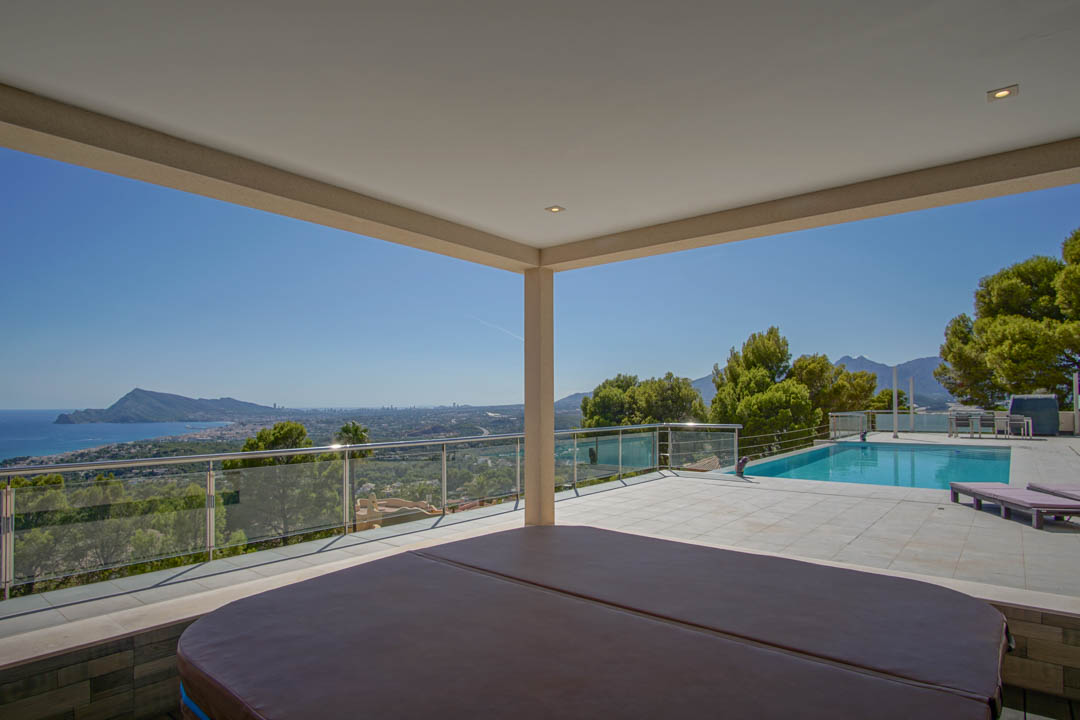 Terraza cubierta con jacuzzi y vistas panorámicas hacia la bahía de Altea y Benidorm, junto a una piscina exterior rodeada de pinos mediterráneos.