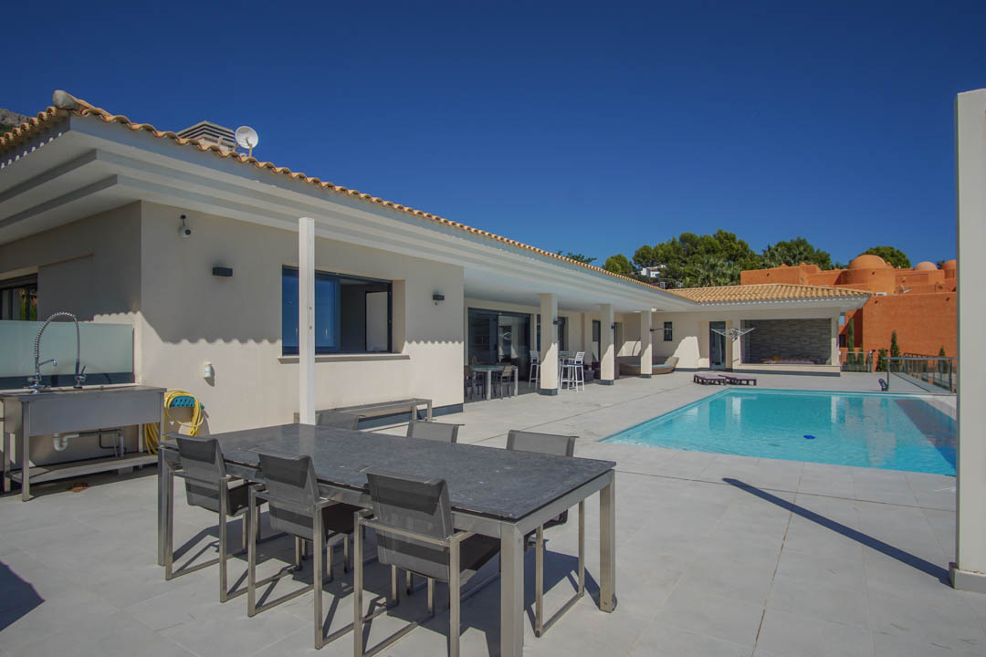 Terraza exterior con piscina rectangular, zona de comedor con mesa de piedra y fregadero industrial de acero inoxidable bajo un cielo despejado.