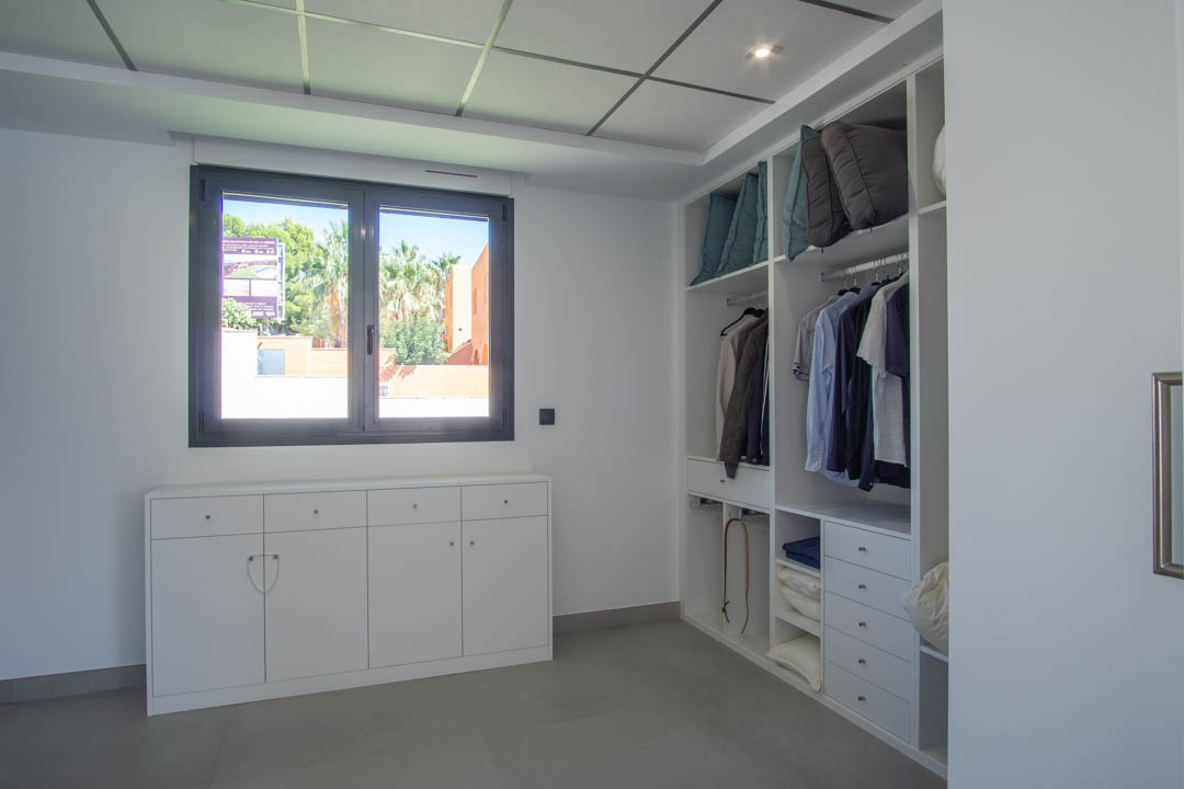 Vestidor con armarios blancos abiertos, cajonera baja bajo la ventana y techo técnico con iluminación integrada sobre suelos de gres gris.