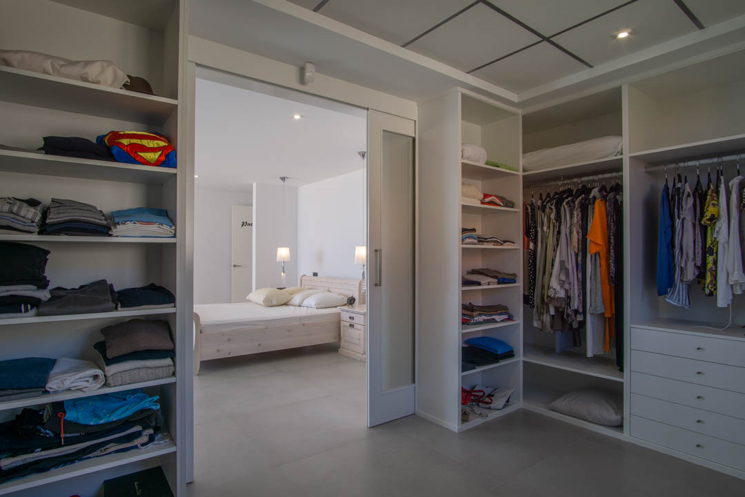 Vestidor de 12m2 con estanterías modulares blancas, cajonera y acceso directo al dormitorio principal mediante una puerta corredera de cristal translúcido.