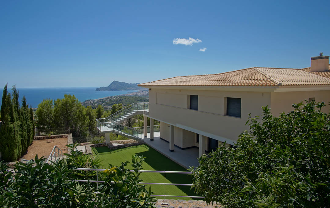 Vista exterior de la villa con jardín de césped artificial, árboles frutales y escalera de cristal, orientada hacia la bahía de Altea y el mar Mediterráneo.
