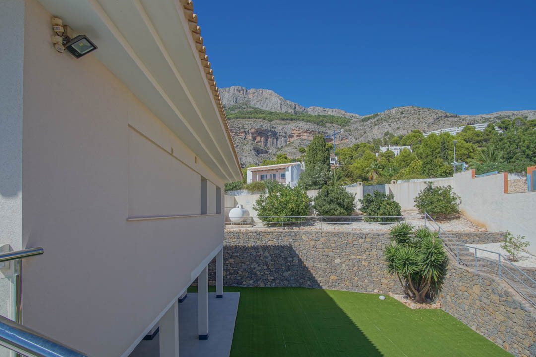 Vista exterior del jardín inferior con césped artificial, muros de piedra natural y escaleras metálicas, con las montañas de la Sierra de Altea al fondo.