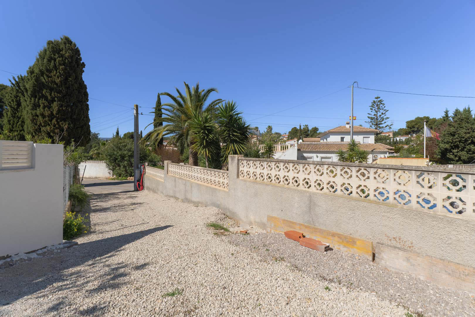 Acceso exterior a la propiedad con suelo de grava blanca y muro perimetral de bloque decorativo, rodeado de vegetación mediterránea y palmeras.
