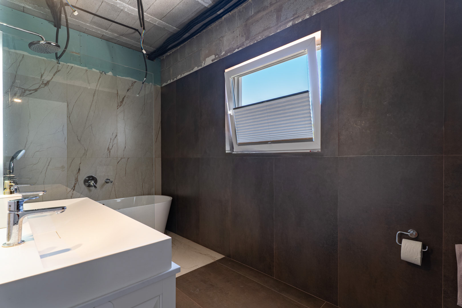 Baño en fase de acabado con lavabo doble, bañera exenta y zona de ducha con revestimiento efecto mármol. Destacan los azulejos de gran formato en tonos oscuros y la ventana oscilobatiente.