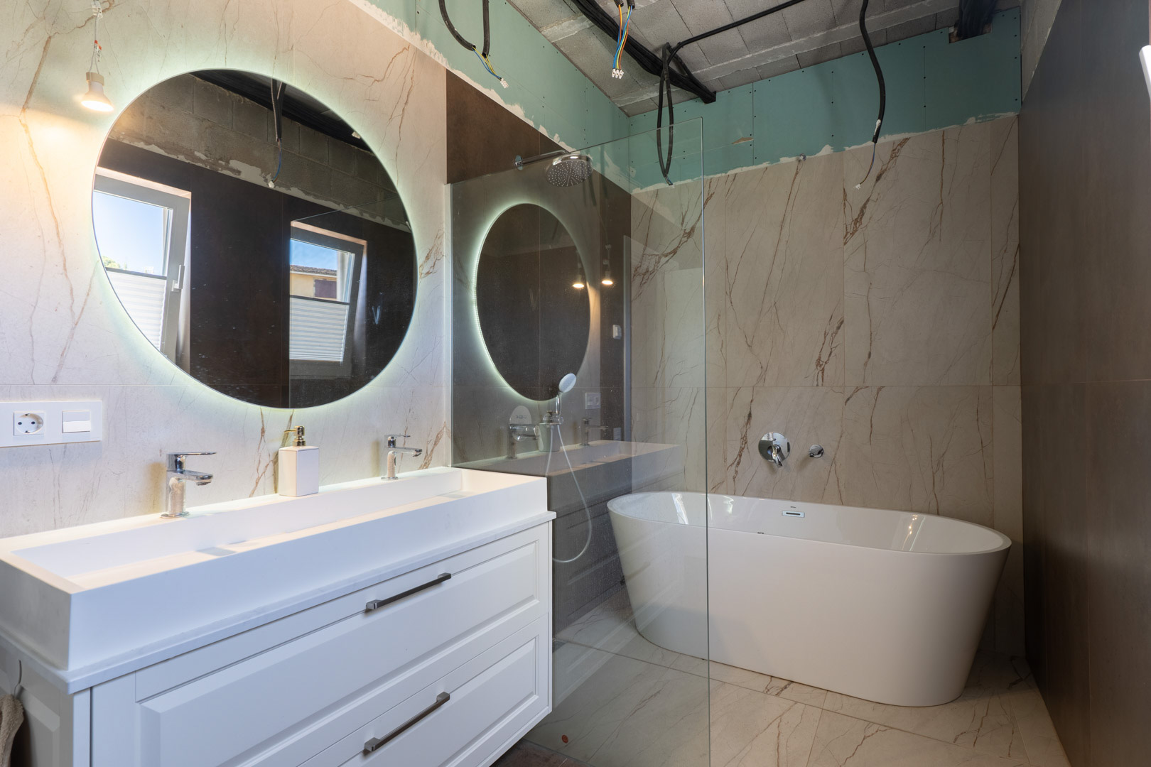 Baño en proceso de finalización con lavabo doble, espejo circular retroiluminado y bañera exenta sobre gres porcelánico con efecto mármol.