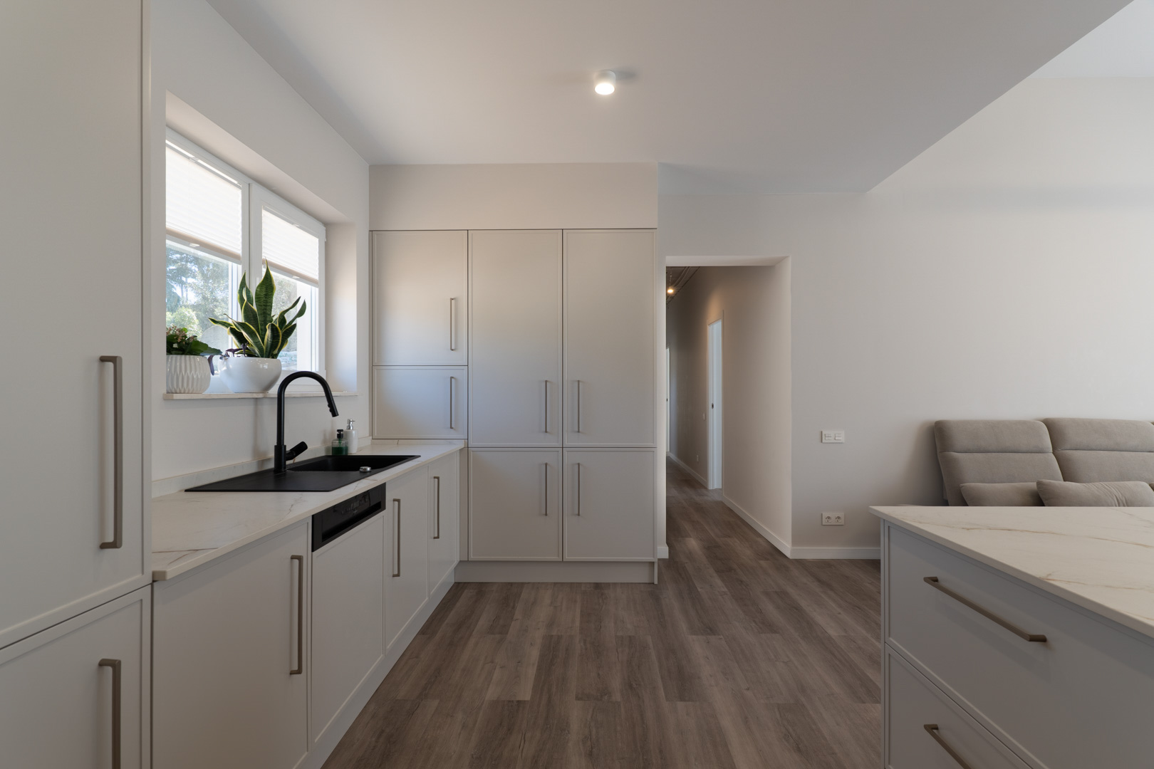 Cocina minimalista con mobiliario en blanco mate, encimera de piedra veteada y grifería negra. El diseño funcional integra suelos de madera y luz natural desde el ventanal.