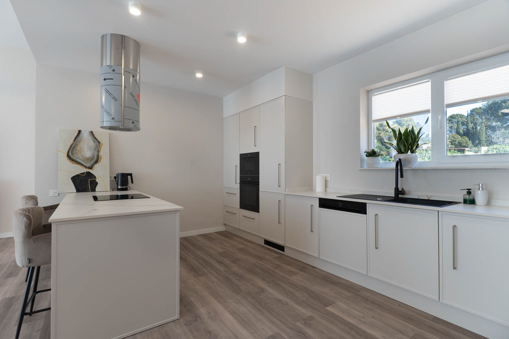Cocina moderna con isla central, encimera blanca y campana cilíndrica. Dispone de electrodomésticos integrados, fregadero negro y suelos de madera laminada bajo luz natural.