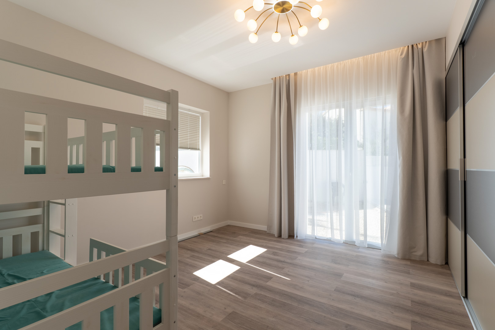 Dormitorio luminoso con literas de madera blanca, suelos laminados y un amplio armario empotrado. La luz natural entra a través de grandes ventanales con cortinas traslúcidas.