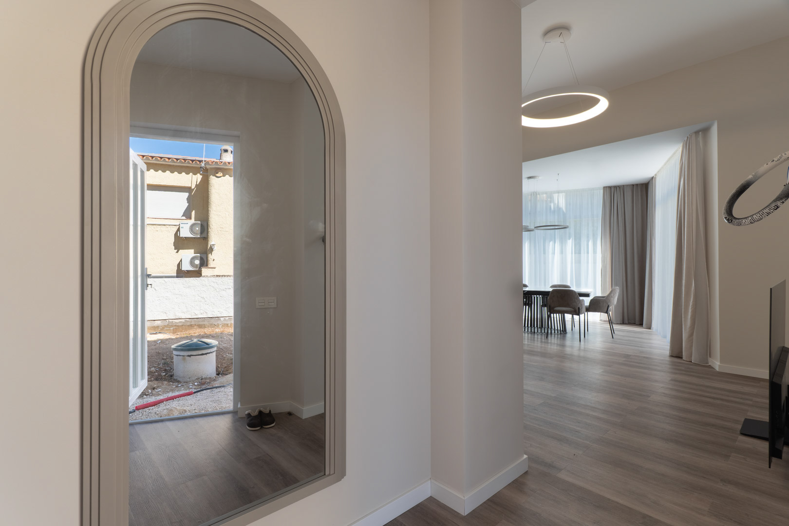 Entrada con espejo de arco que refleja el exterior, conectando con un salón de diseño minimalista, suelos laminados y luminaria circular LED.