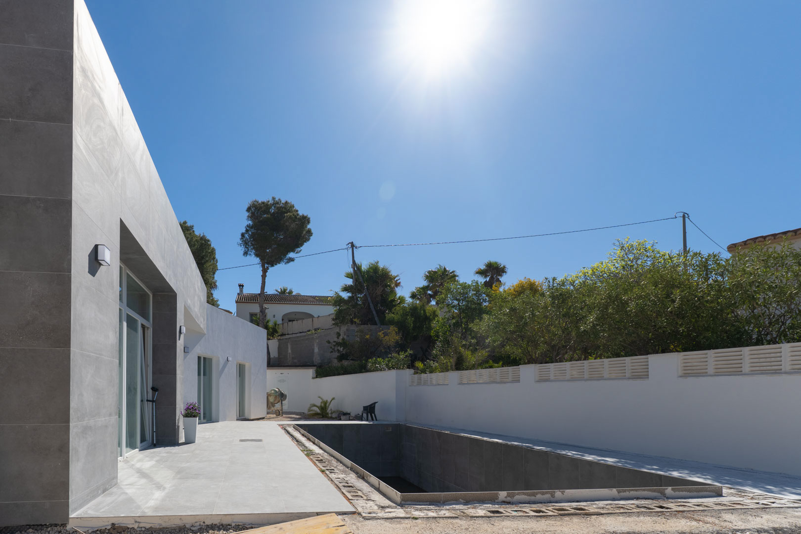Fachada minimalista con aplacado gris y zona de piscina en proceso de terminación, rodeada de un muro perimetral blanco bajo el sol mediterráneo.