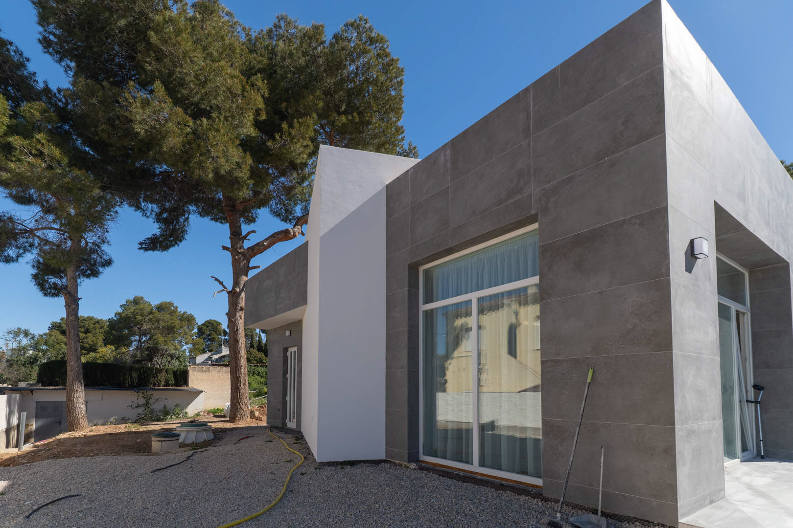 Fachada minimalista con revestimiento cerámico gris y grandes ventanales, rodeada de pinos maduros y zonas exteriores actualmente en fase de acondicionamiento.