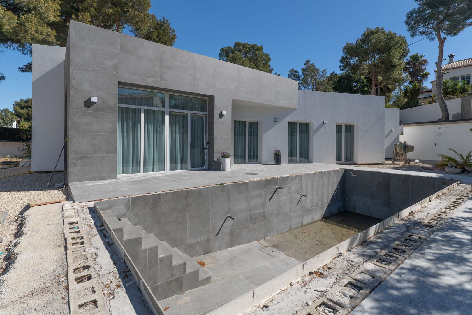 Fachada minimalista de una planta con revestimiento porcelánico gris y piscina en proceso de construcción, rodeada de pinos mediterráneos.