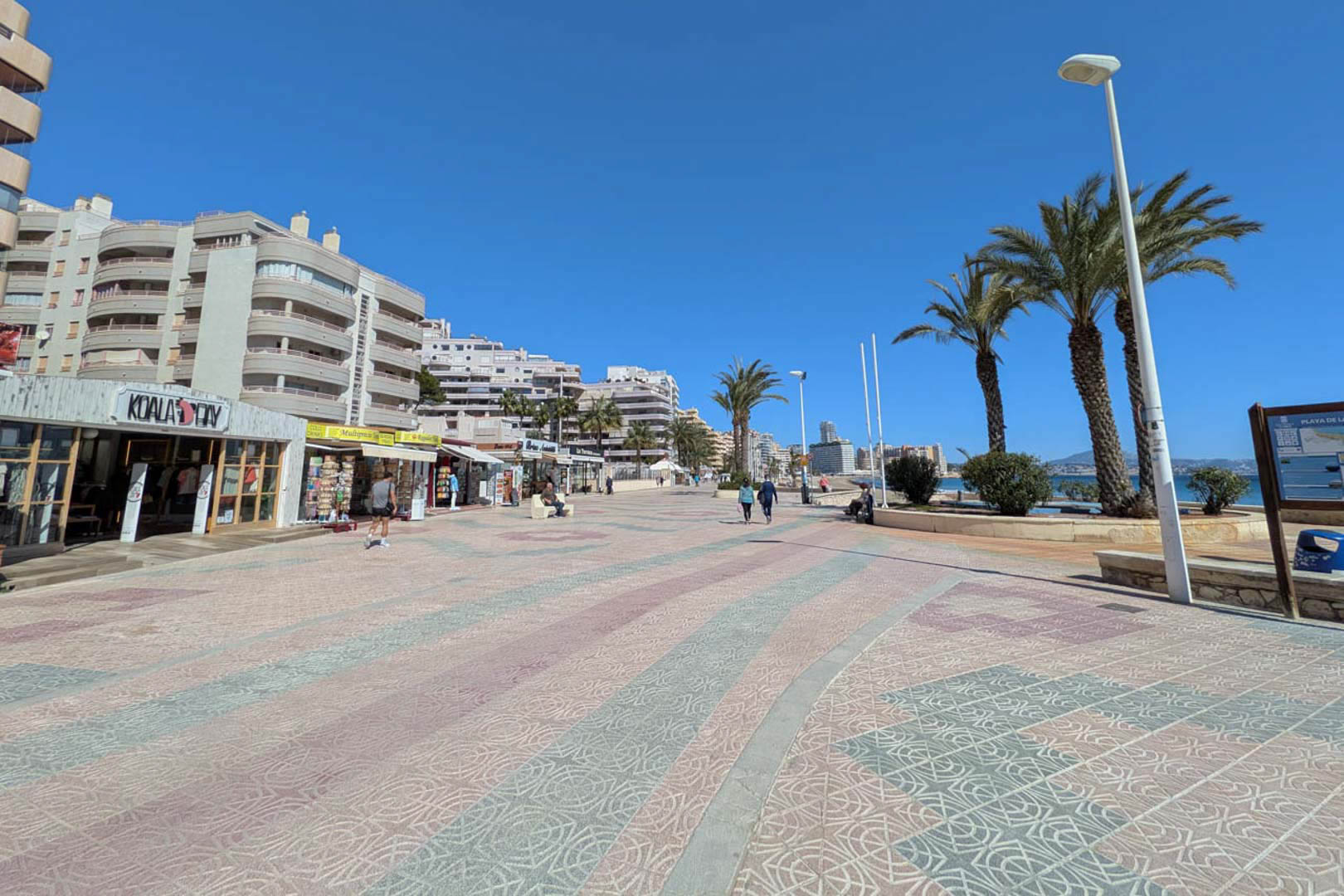 Paseo marítimo de Calpe con pavimento decorativo, rodeado de comercios locales, palmeras y edificios residenciales bajo un cielo despejado.