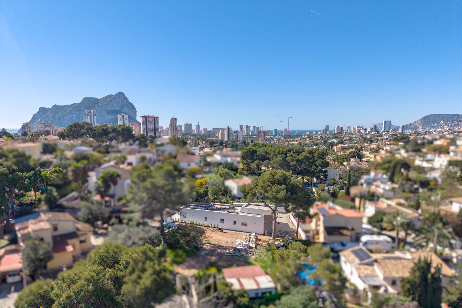 Vista aérea de la villa minimalista con el Peñón de Ifach al fondo. Se aprecia la estructura de una planta y la parcela en fase de terminación exterior.