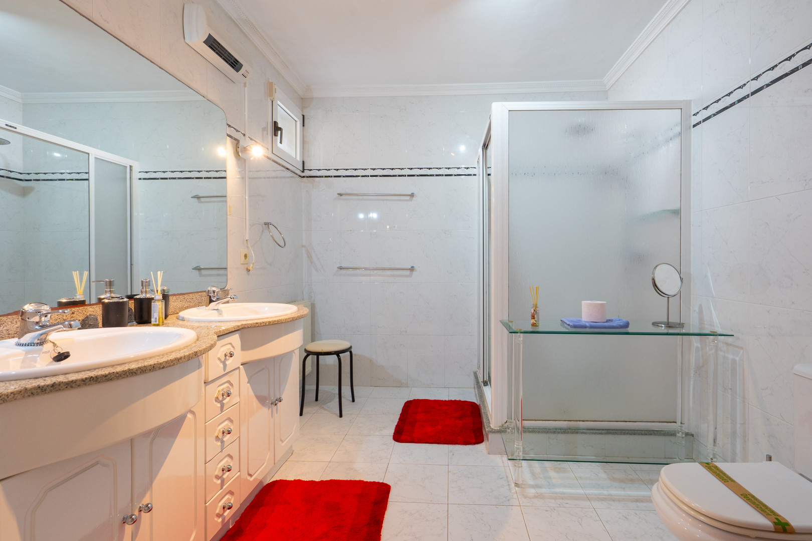 Baño con tocador de doble seno, encimera de granito y armarios blancos. Incluye una amplia ducha con mampara translúcida y aire acondicionado individual.