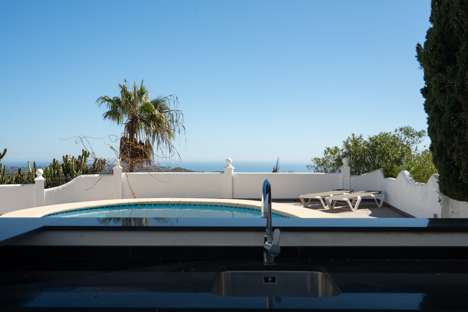 Cocina exterior con encimera oscura y fregadero, con vistas a la piscina circular y al mar Mediterráneo bajo un cielo despejado.