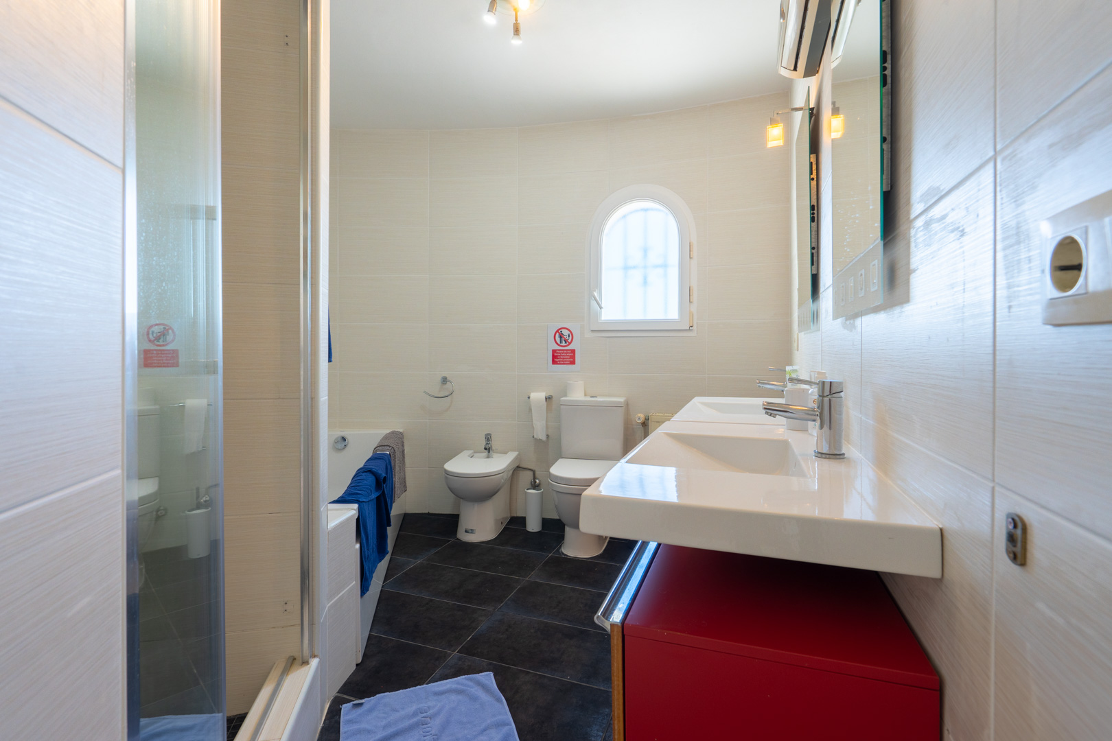 Cuarto de baño con lavabo doble sobre mueble rojo, inodoro, bidé y bañera. Paredes alicatadas en tonos claros, suelo oscuro y una pequeña ventana de arco.