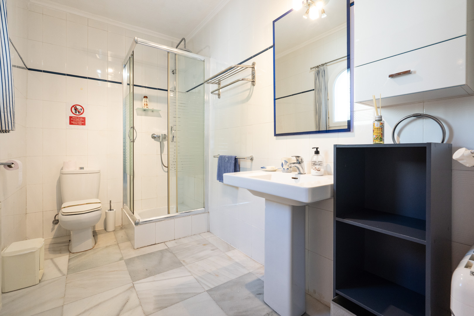 Cuarto de baño con paredes alicatadas en blanco, suelo de mármol, cabina de ducha de cristal y lavabo de pedestal con espejo de marco azul.