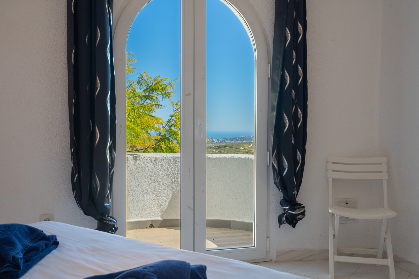 Dormitorio con puerta de arco acristalada que ofrece vistas al mar y acceso a un balcón privado. Presenta paredes blancas y cortinas oscuras con motivos.