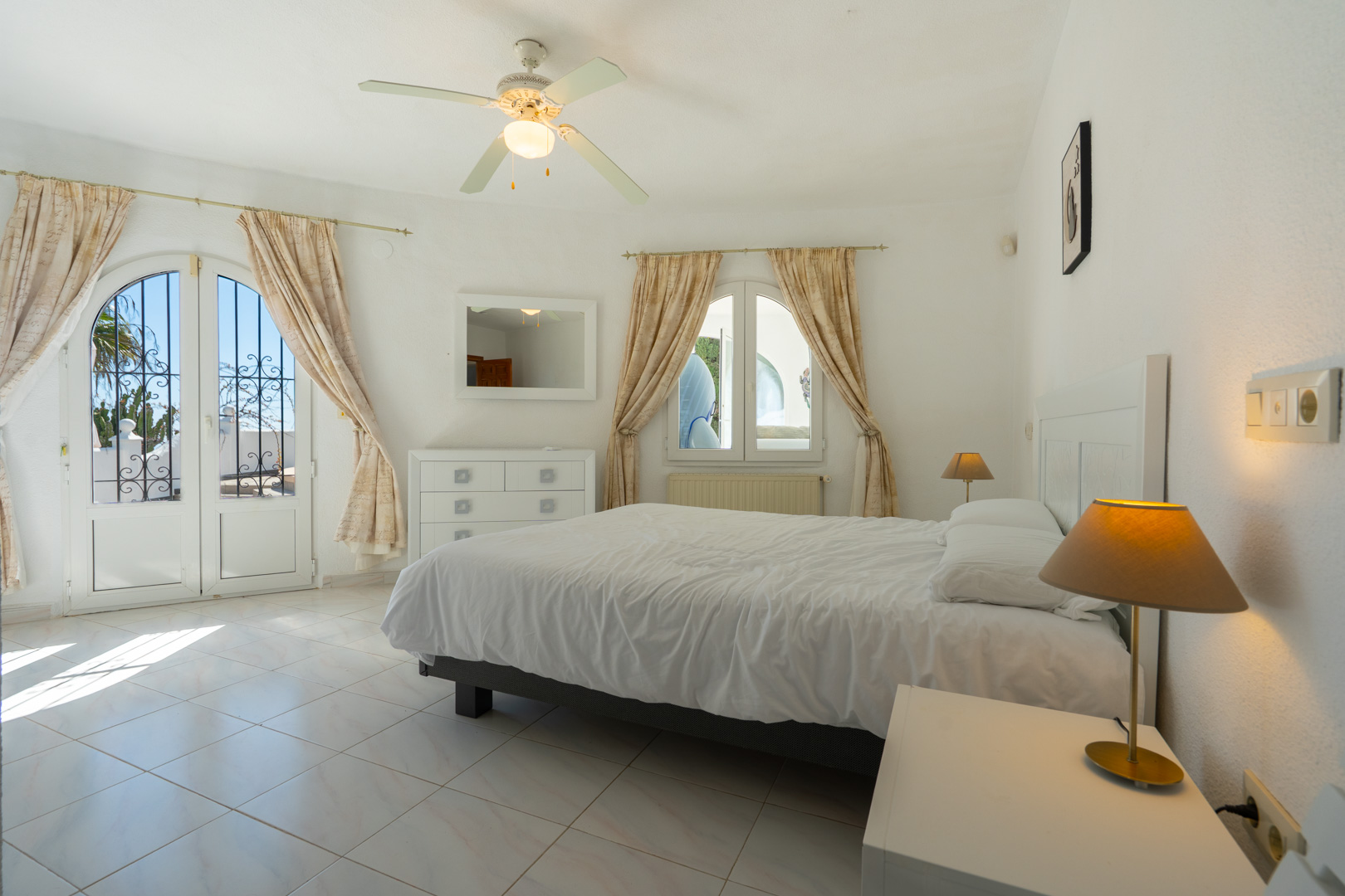 Dormitorio luminoso con suelos de gres, ventilador de techo y acceso directo al exterior a través de una puerta acristalada con arco y rejería decorativa.