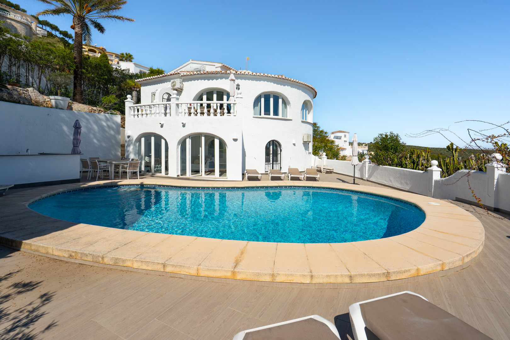 Exterior de la villa con piscina curva, amplia terraza de gres porcelánico imitación madera y fachada blanca con arcos y balaustrada tradicional.