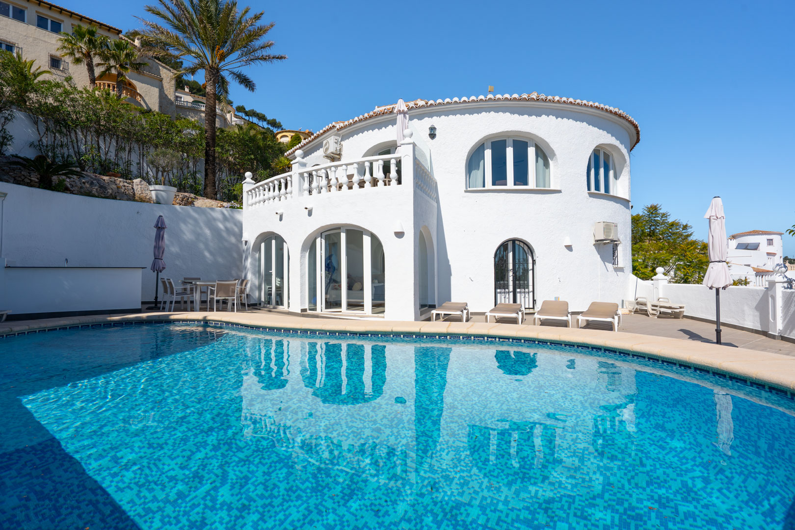 Fachada mediterránea de la villa con piscina privada, naya acristalada y terraza solárium. Destacan los ventanales en arco y la balaustrada blanca tradicional.