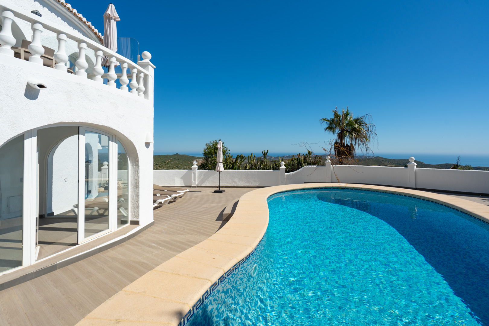Piscina de forma curva con bordes cerámicos y amplia terraza de gres efecto madera, con vistas panorámicas al mar y acceso directo a la naya acristalada.