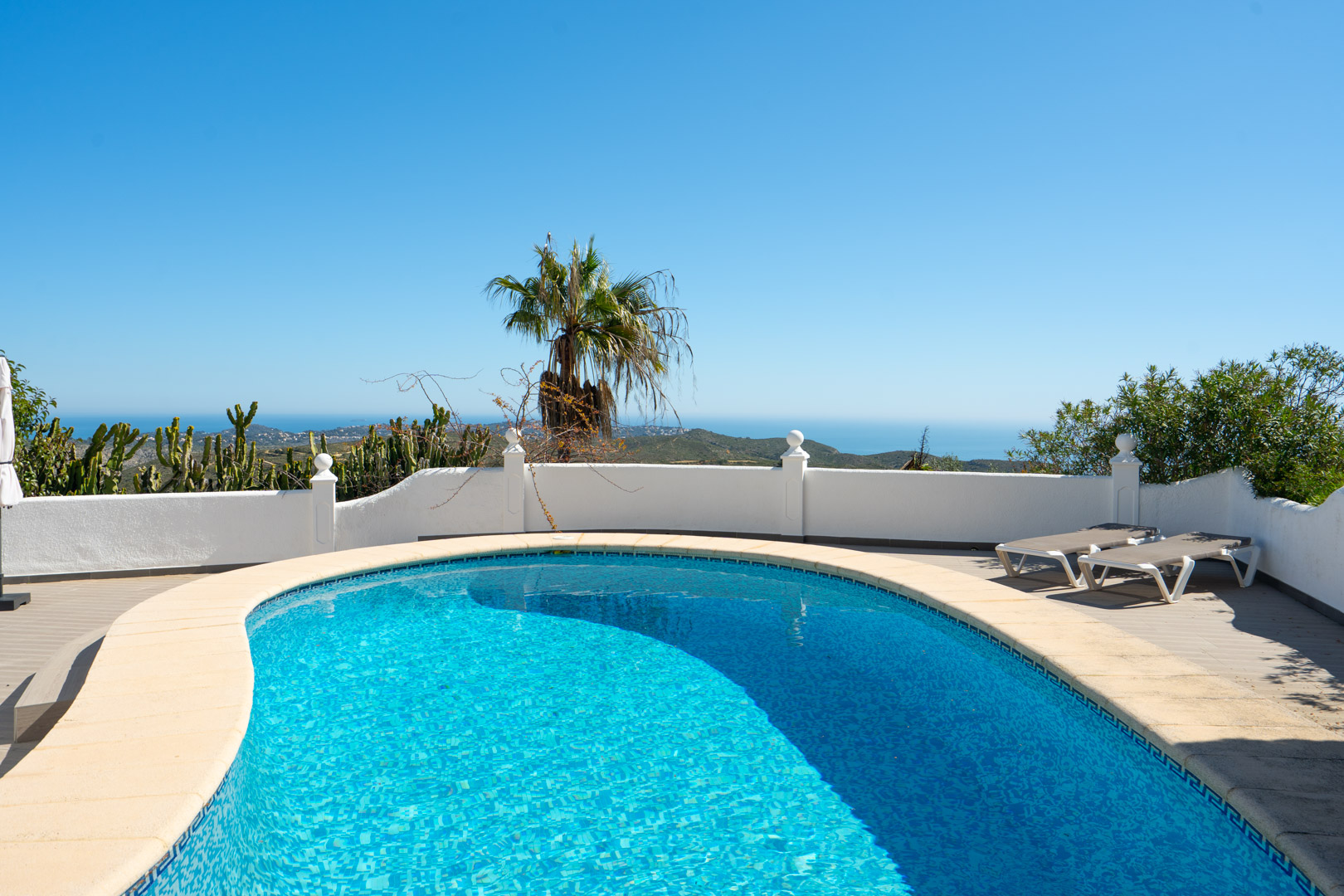 Piscina de forma curva con vistas panorámicas al mar Mediterráneo, rodeada por una terraza solárium con tumbonas y muros blancos de estilo tradicional.