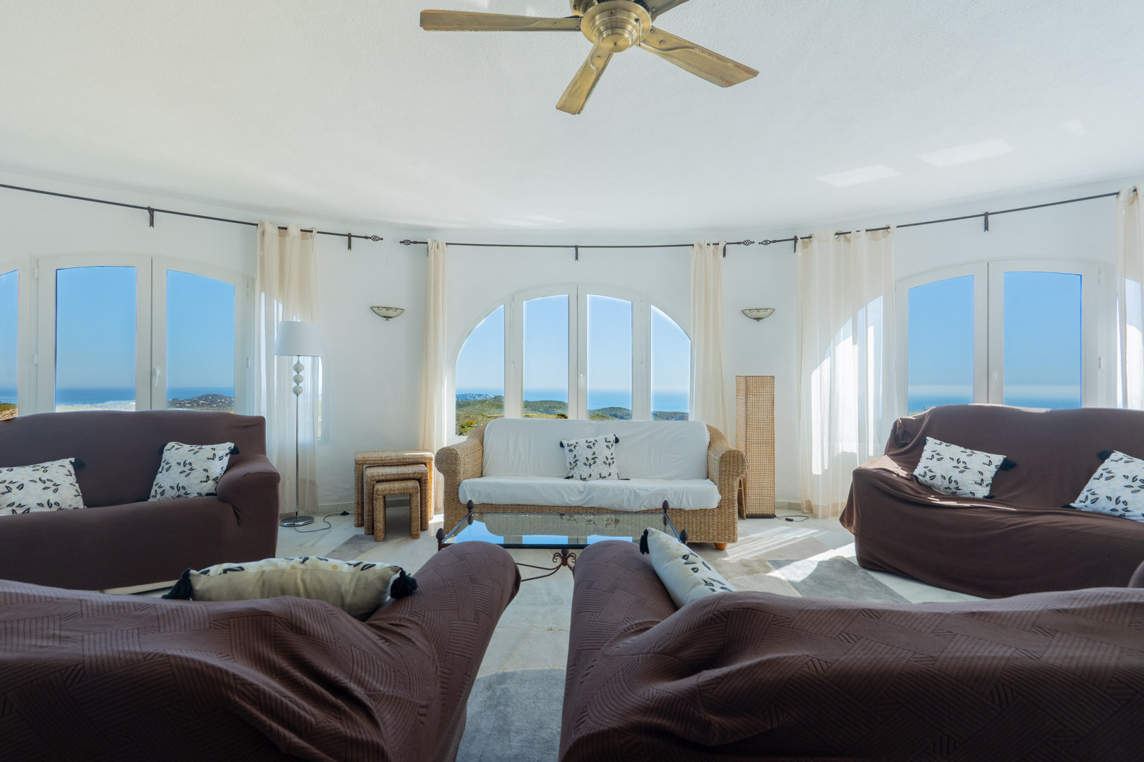 Salón circular con amplios ventanales en arco que ofrecen vistas panorámicas al mar. Incluye mobiliario confortable, ventilador de techo y abundante luz natural.